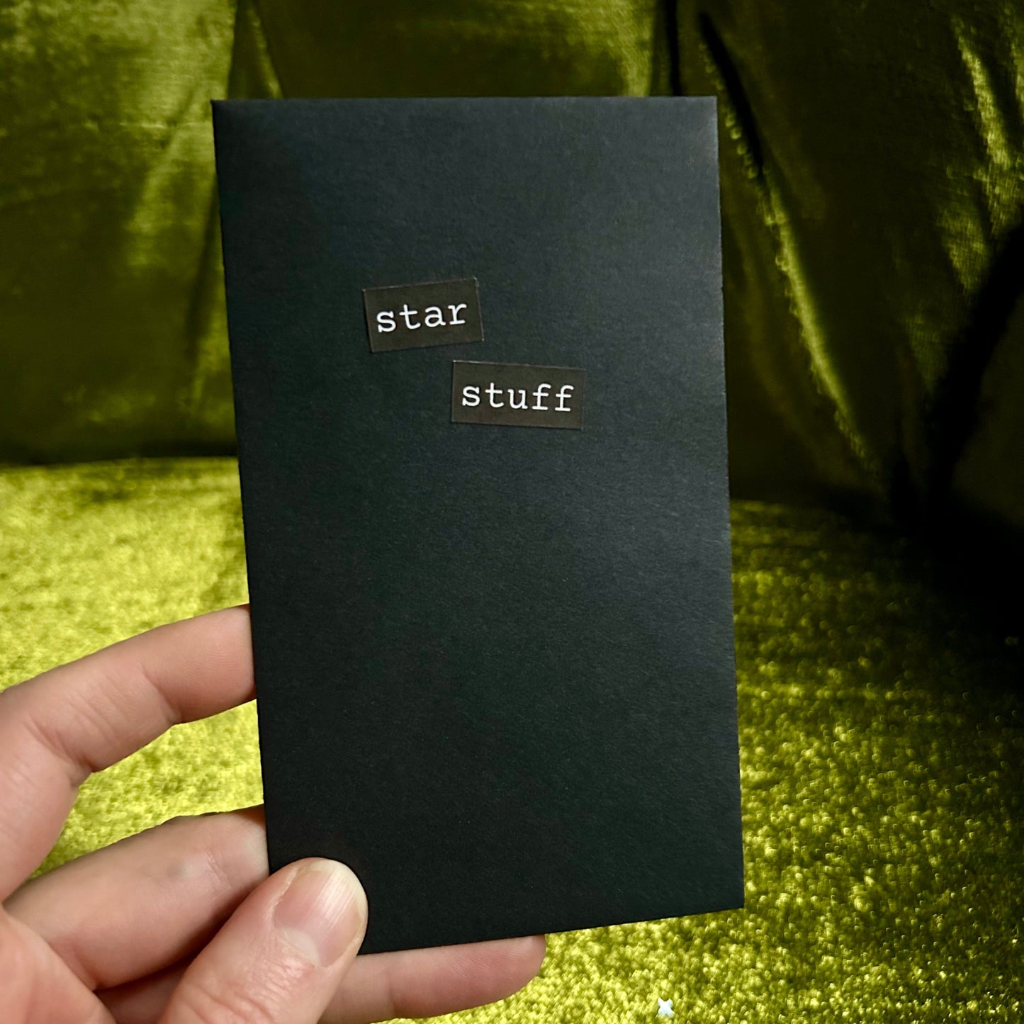 Black envelope with 'star stuff' title held by a hand against a green glittery background