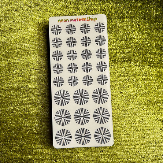 Sheet of black circular stickers on a white card with 'neon mothership' text, placed on a textured green surface.