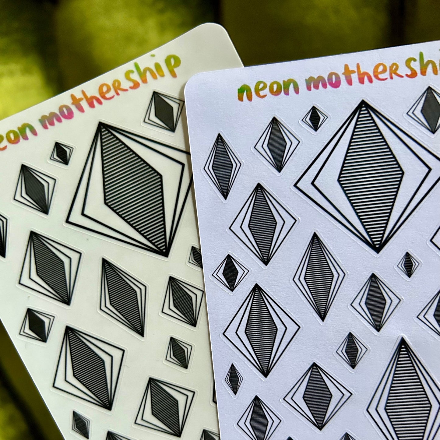 Two sticker sheets with geometric diamond pattern and 'neon mothership' text on a blurred green background