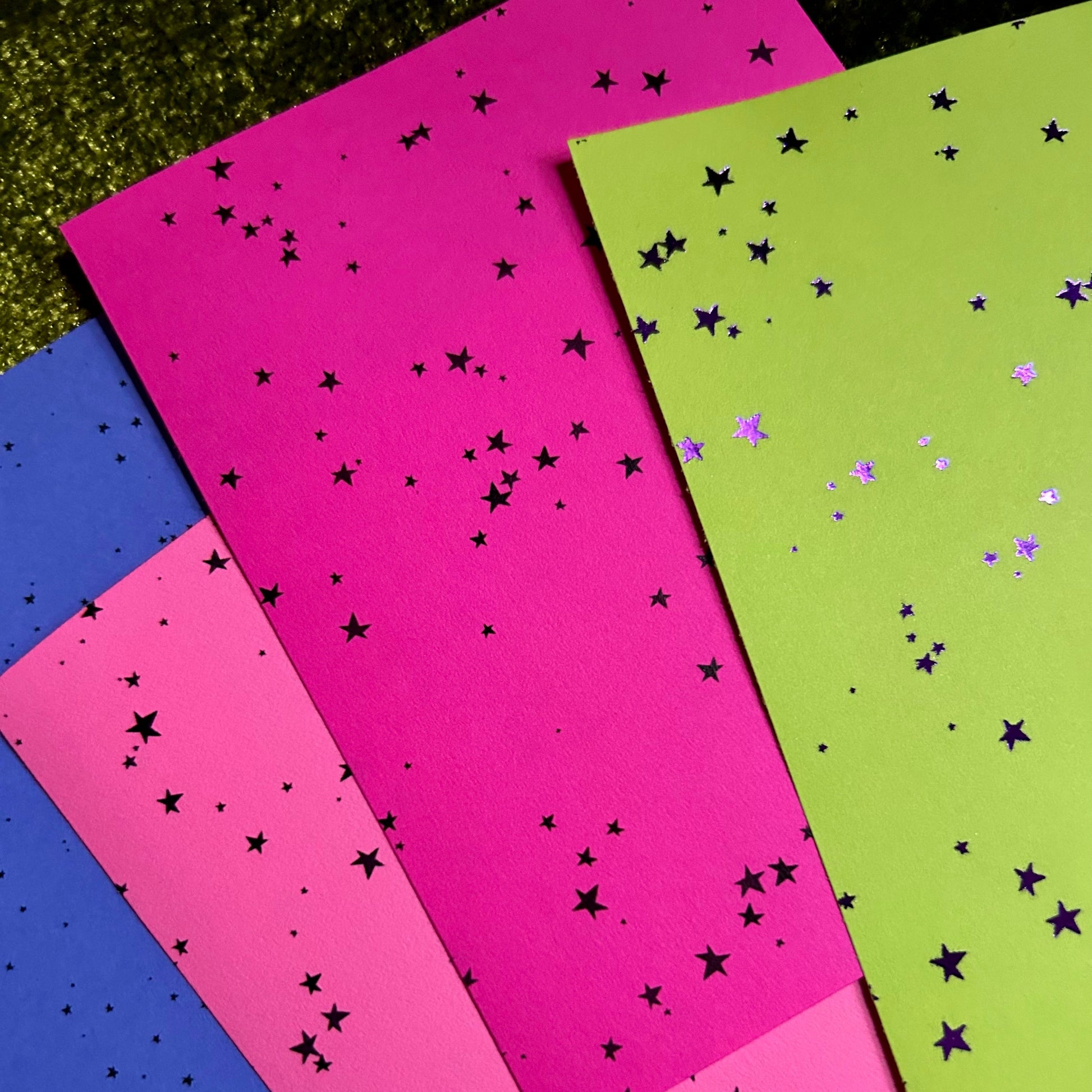 Four sheets of paper in pink, green, and blue with star patterns on a grassy background.