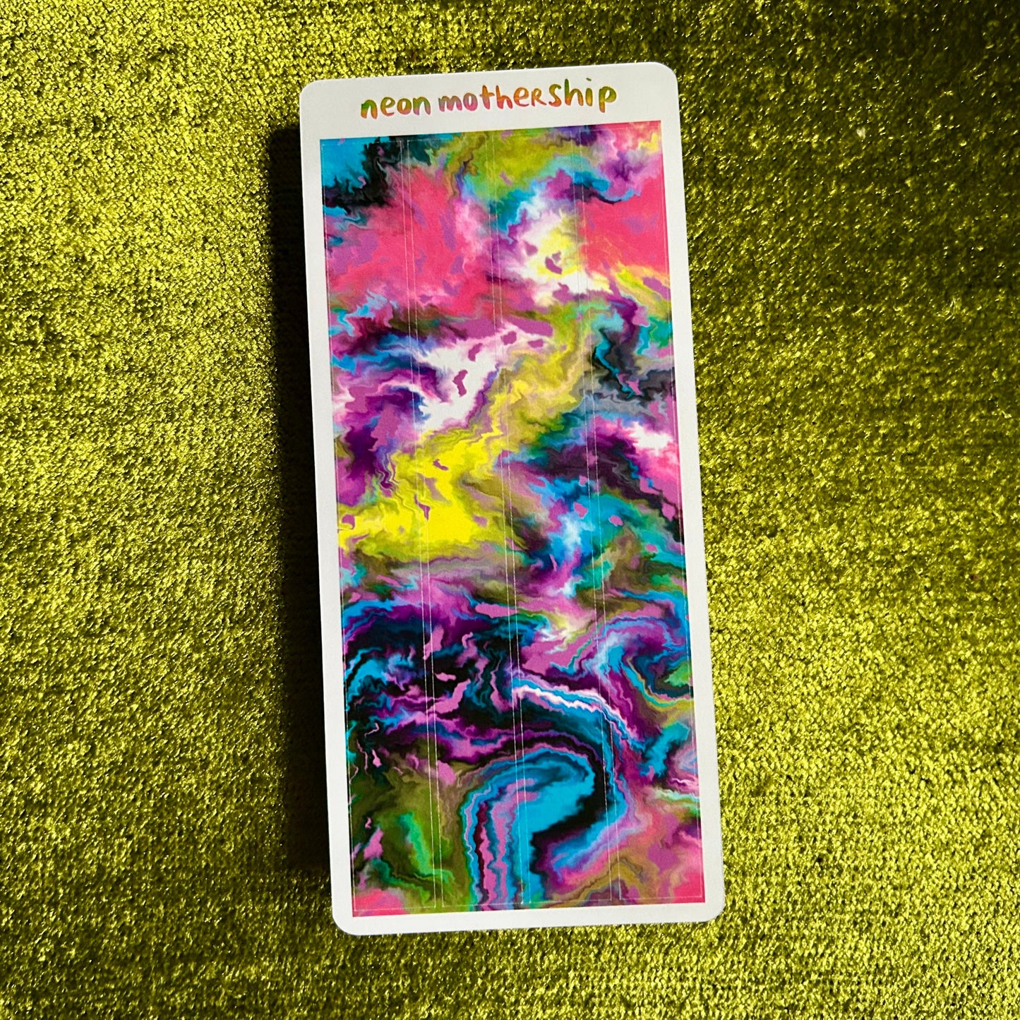 Colorful abstract design on a rectangular card with 'neon mothership' text, placed on a textured green surface.