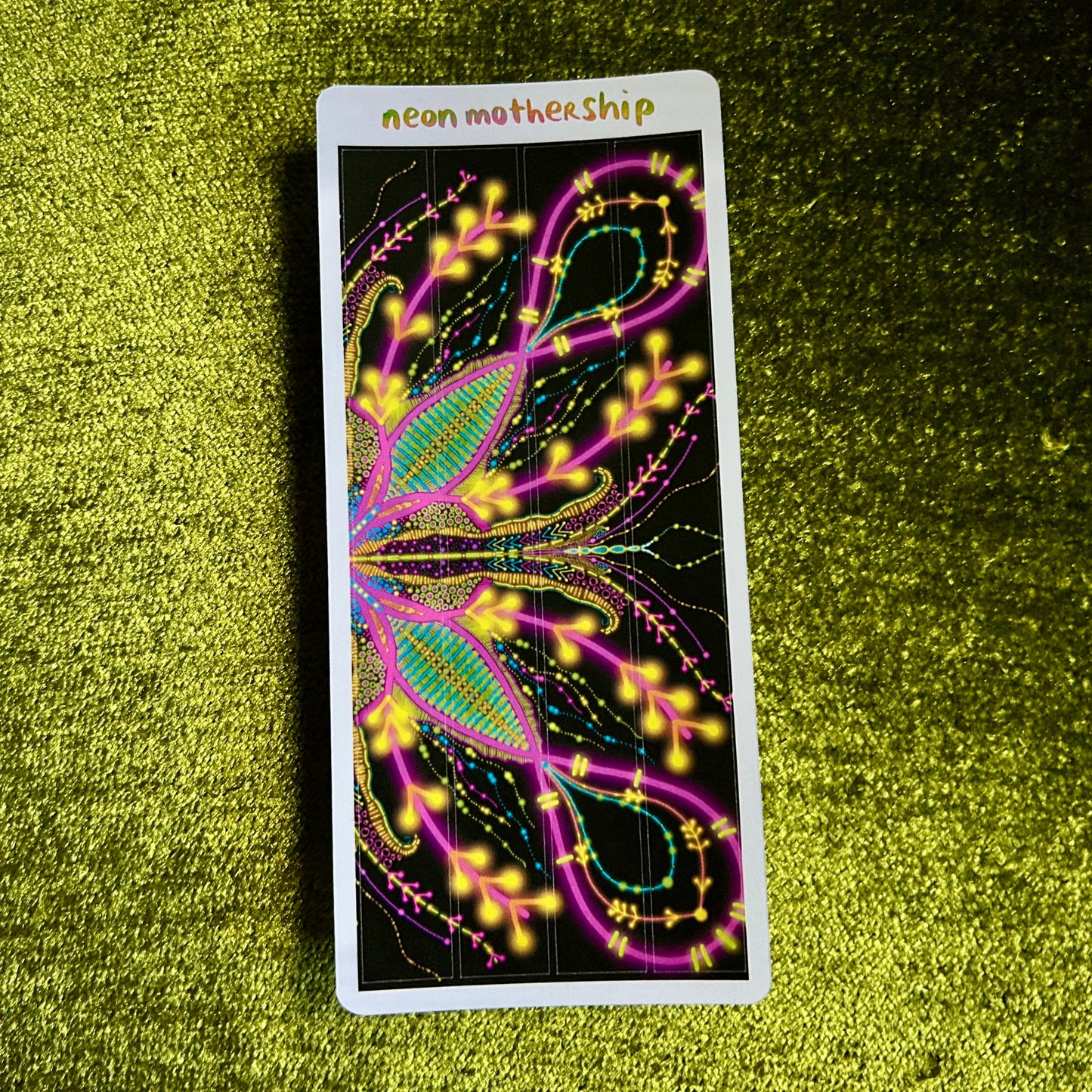 Colorful glowing design on a sticker sheet with 'neon mother ship' branding, placed on a green textured surface.
