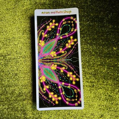 Colorful glowing design on a sticker sheet with 'neon mother ship' branding, placed on a green textured surface.