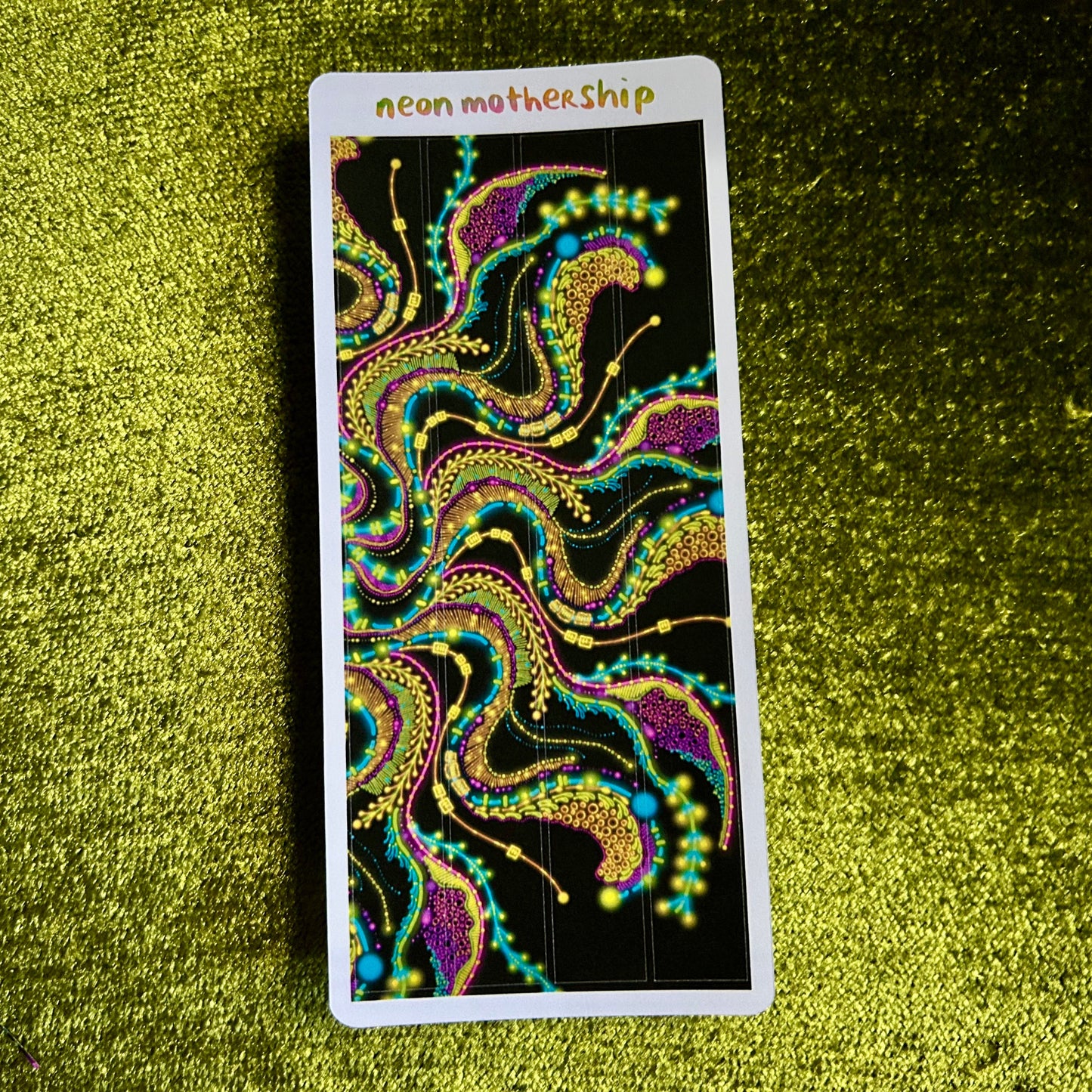 Colorful abstract design on a sticker sheet with 'neon motherShip' text, placed on a green textured surface.