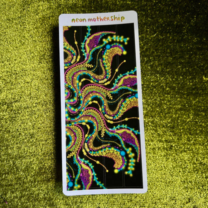 Colorful abstract design on a sticker sheet with 'neon motherShip' text, placed on a green textured surface.