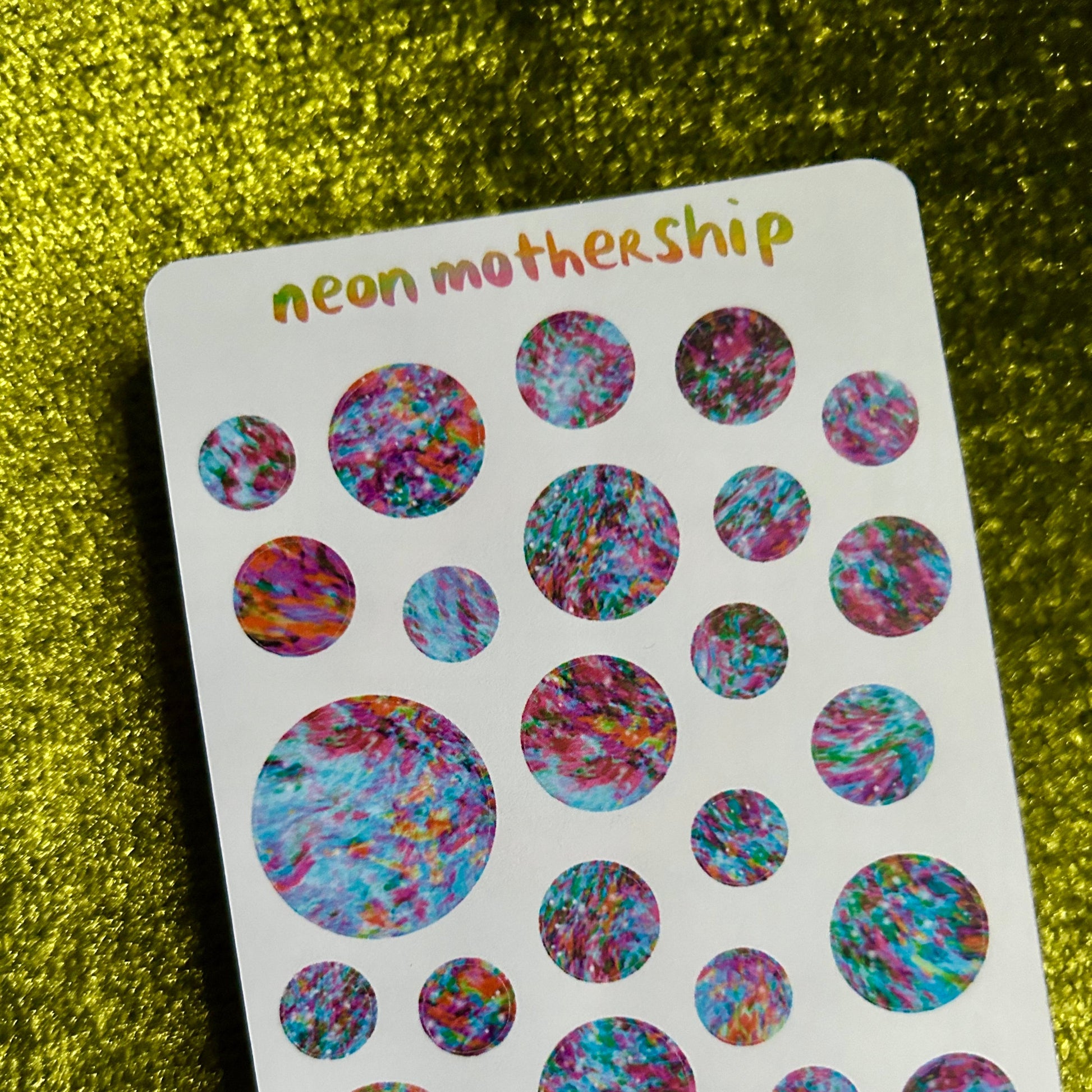 Sheet of colorful round stickers labeled 'neon mothership' on a green textured background