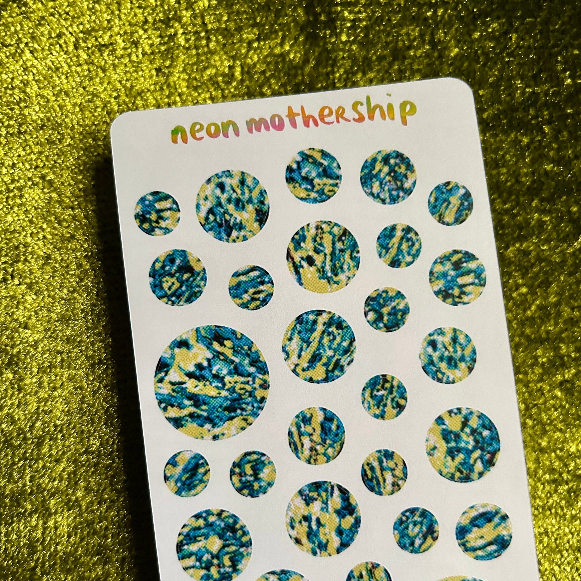 Sheet of round, multicolored stickers with 'neon motherShip' branding on a green textured background.