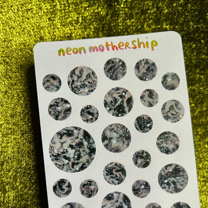 Pack of round stickers with a speckled pattern on a white card labeled 'neon mothership' against a green glittery background.