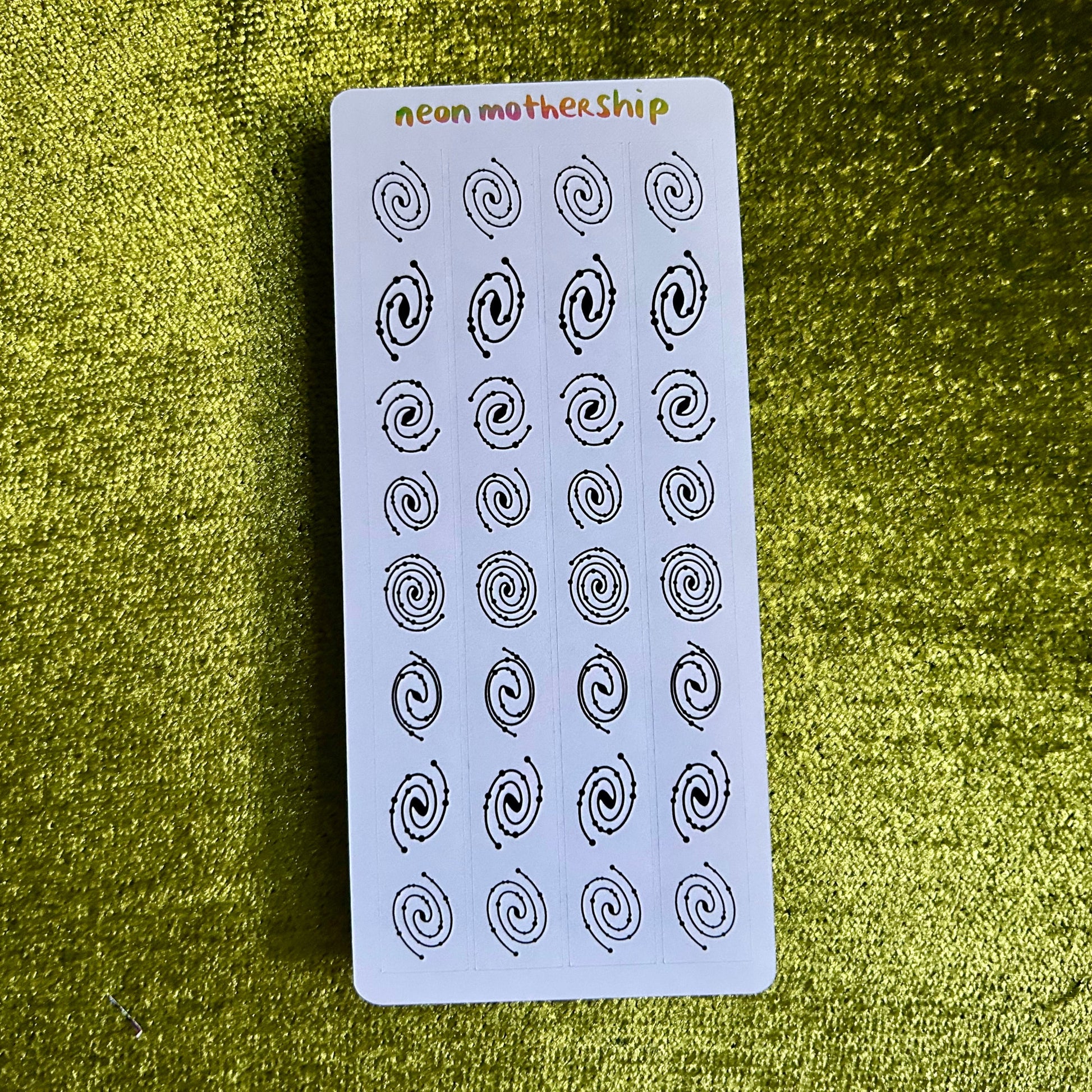 Sheet of black spiral patterns on a white card with 'neon motherhip' branding, placed on a green textured surface.
