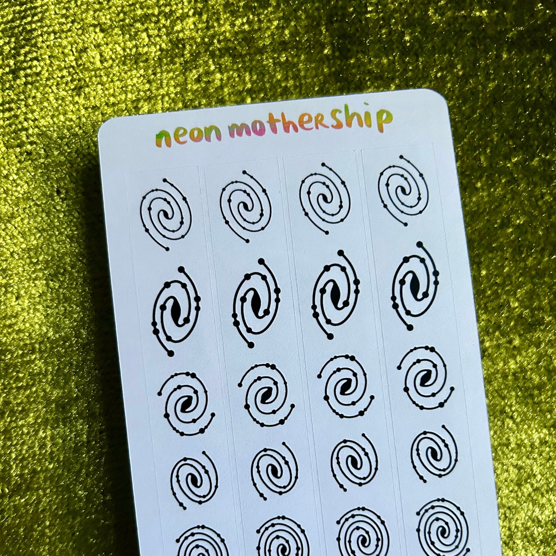 Sheet of black spiral stickers on a white card with 'neon mothership' branding, placed on a green textured surface.