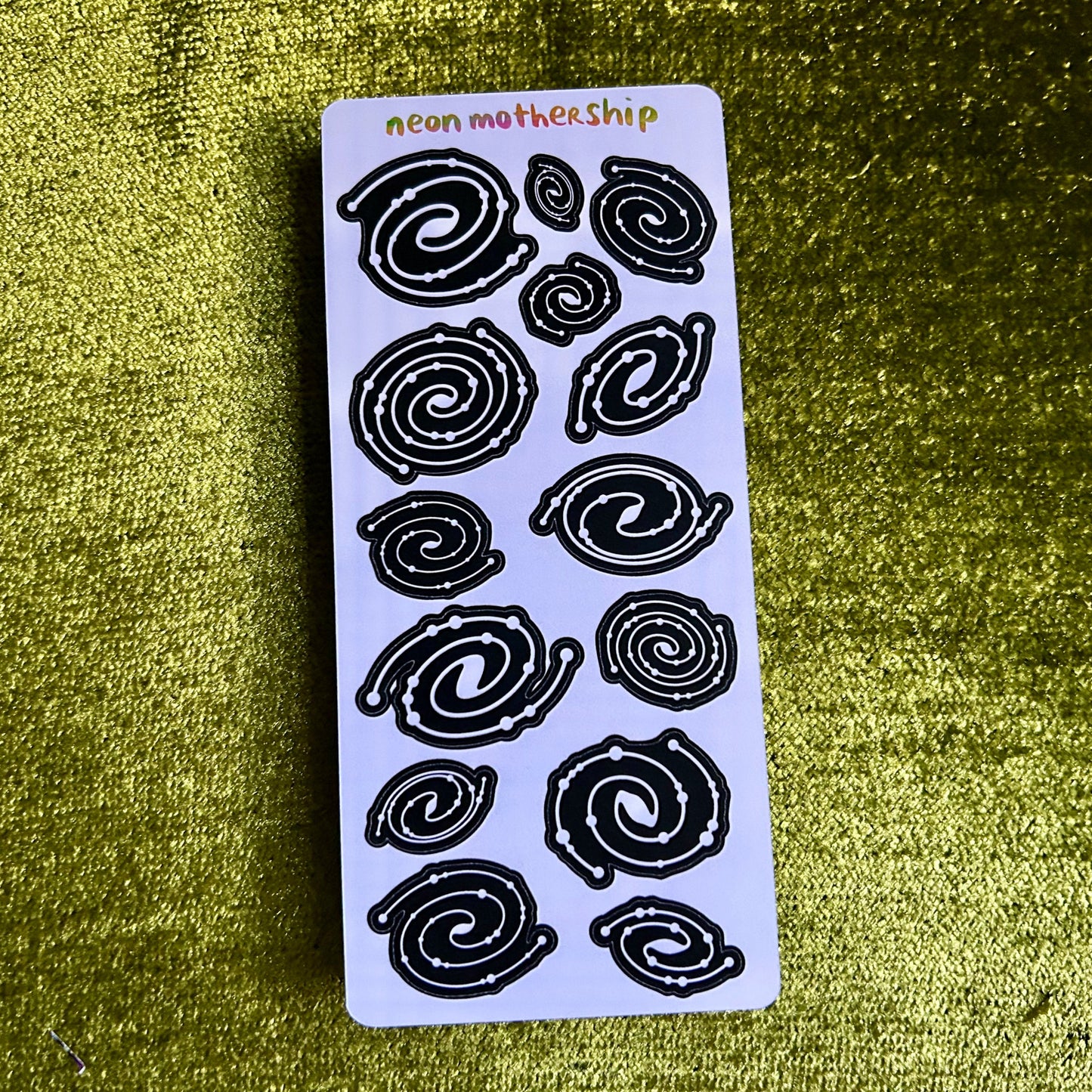 Sheet of black spiral stickers on a white card with 'neon mothership' branding, against a textured green background.