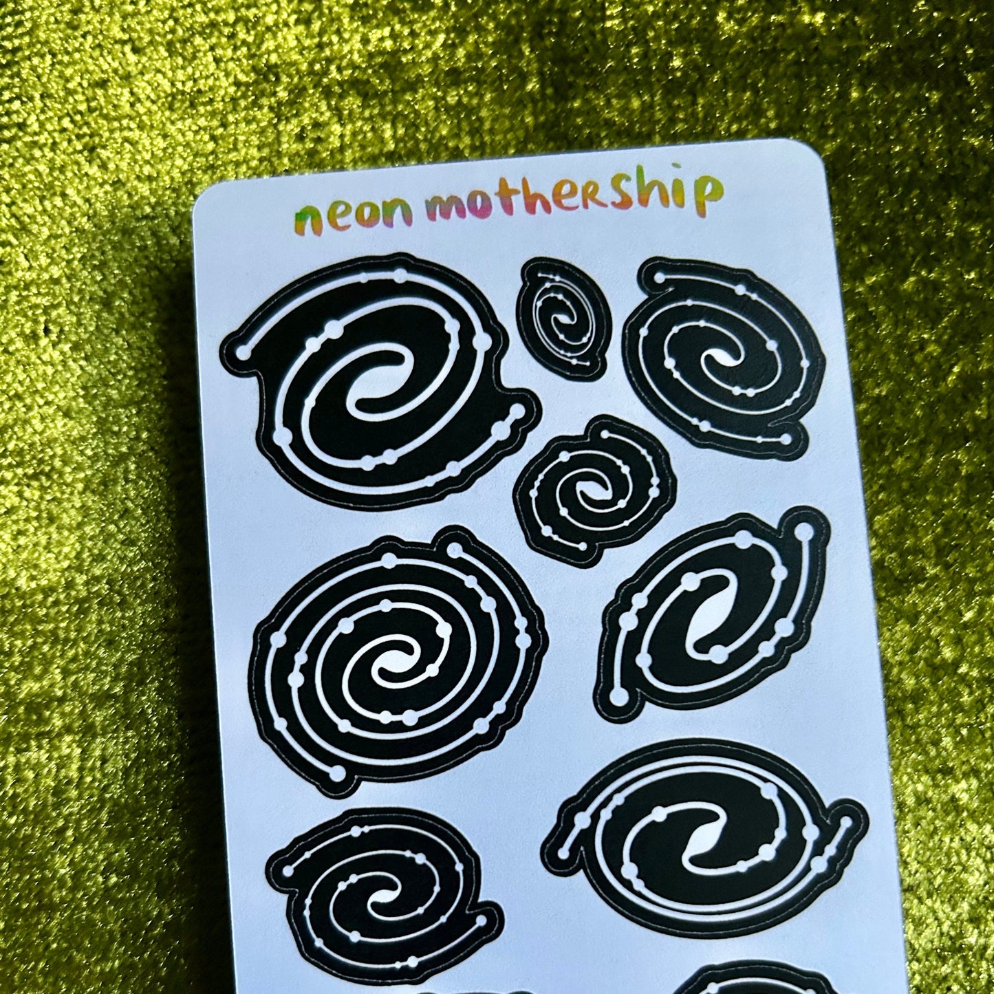 Sheet of black spiral designs on a light blue card with 'neon mothership' text, against a green textured background.