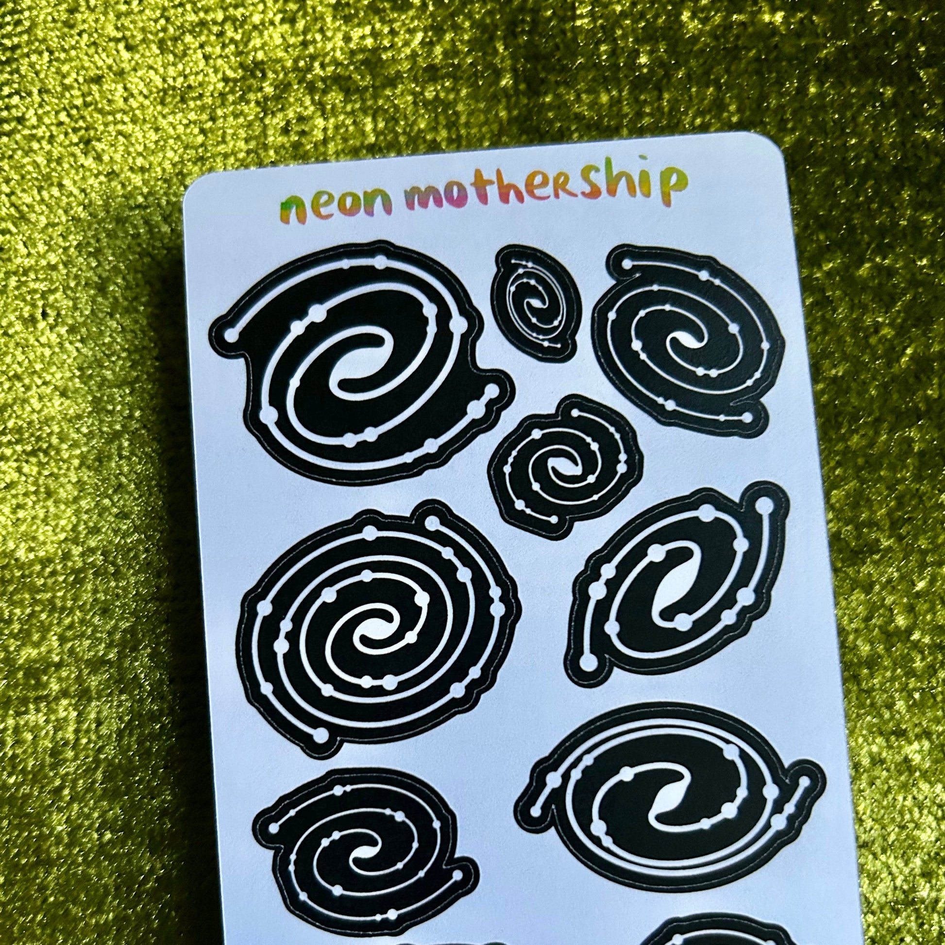 Sheet of black spiral designs on a light blue card with 'neon mothership' text, against a green textured background.
