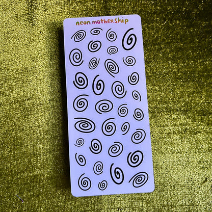 Sheet of black spiral designs on a purple card with 'neon mother ship' branding, on a textured green surface.