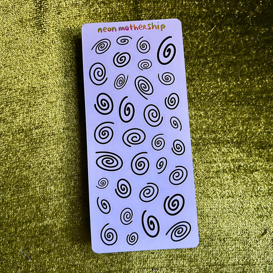 Sheet of black spiral designs on a purple card with 'neon mother ship' branding, on a textured green surface.
