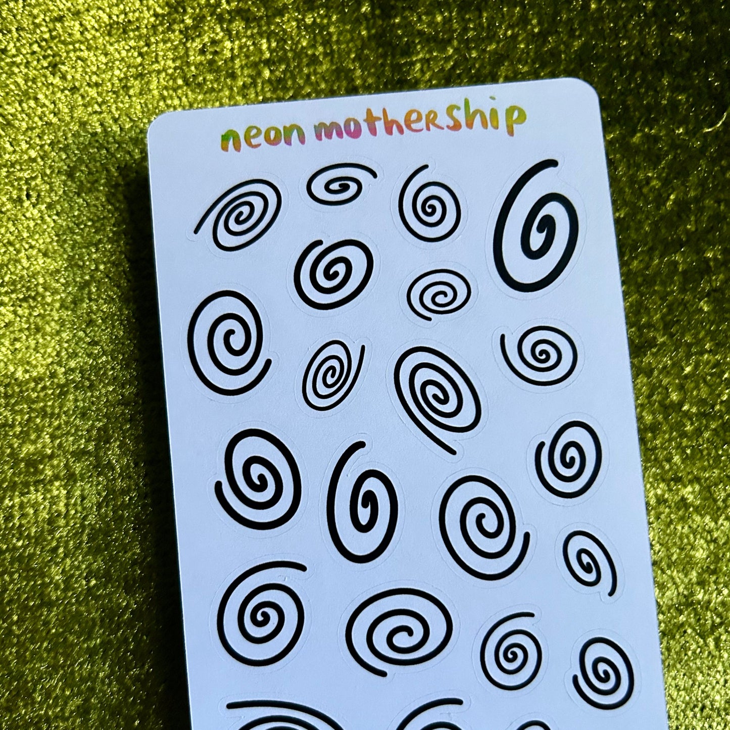 Sheet of black spiral patterns on a blue card with 'neon mothership' branding, against a green textured background.