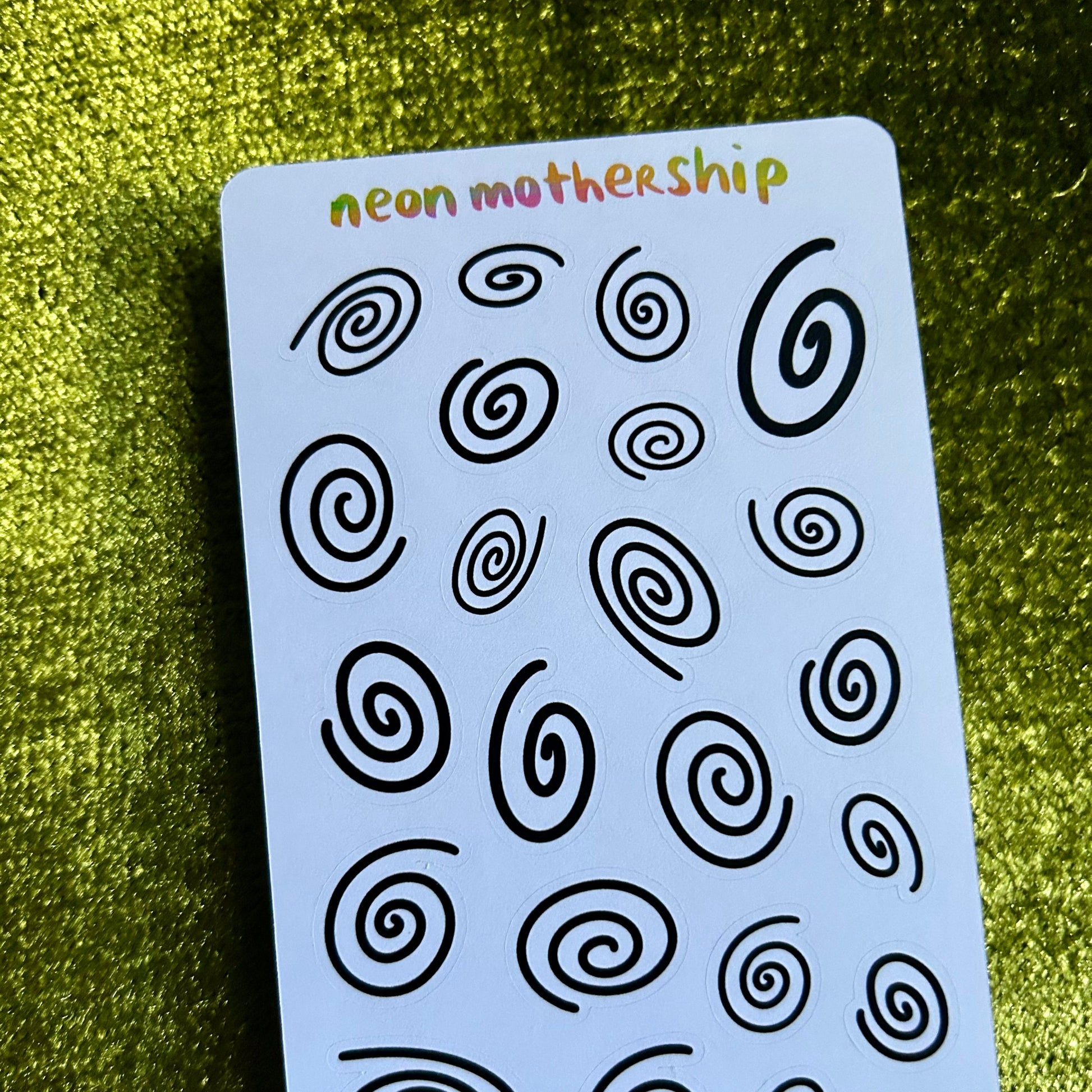Sheet of black spiral patterns on a blue card with 'neon mothership' branding, against a green textured background.