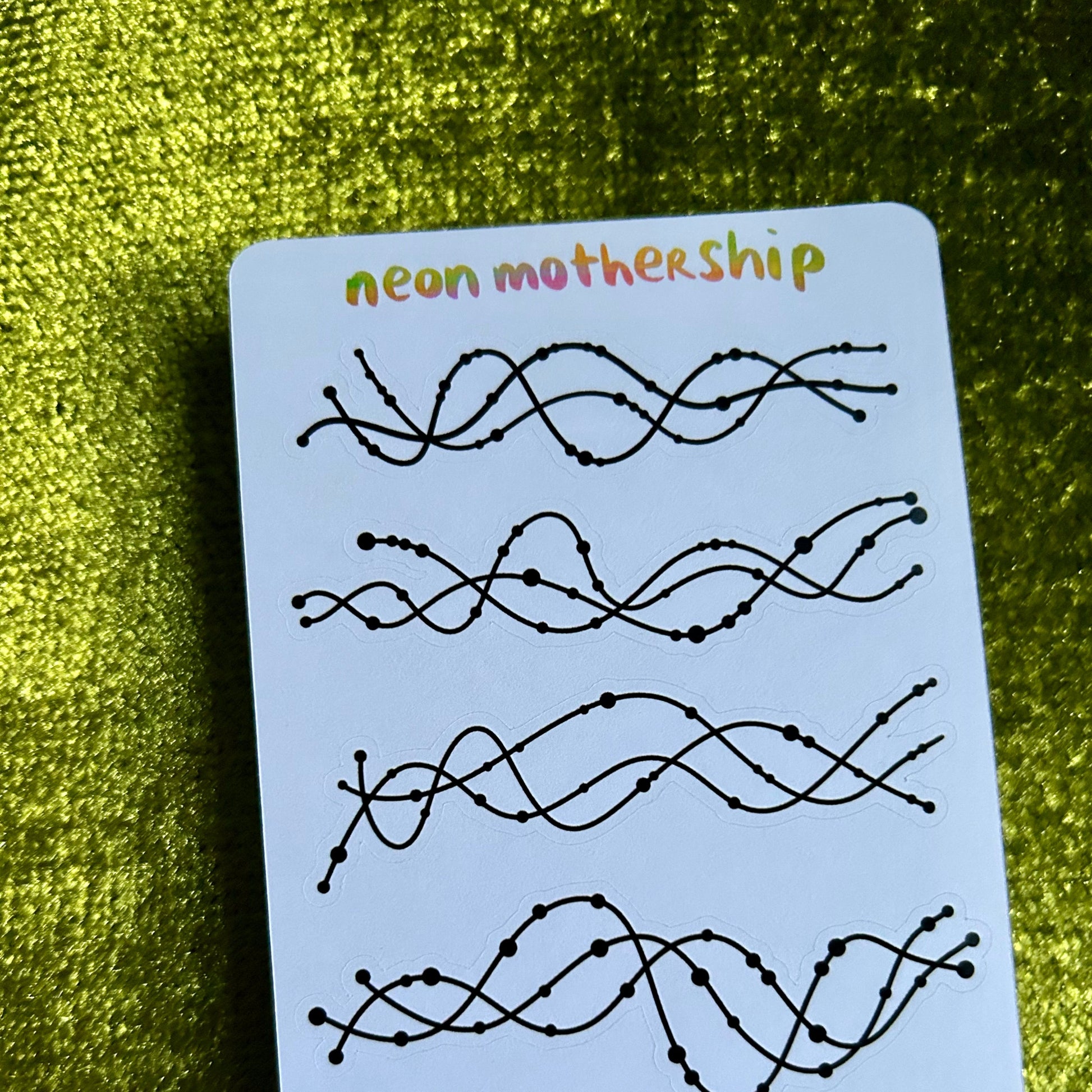 Sheet of black wavy line designs on a white sticker sheet with 'neon mothership' text, against a green textured background.