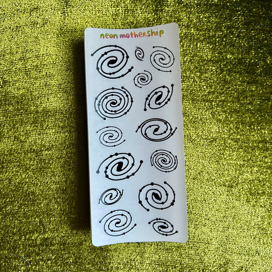 Sheet of black spiral designs on a white card with 'neon mothership' branding, placed on a green textured surface.