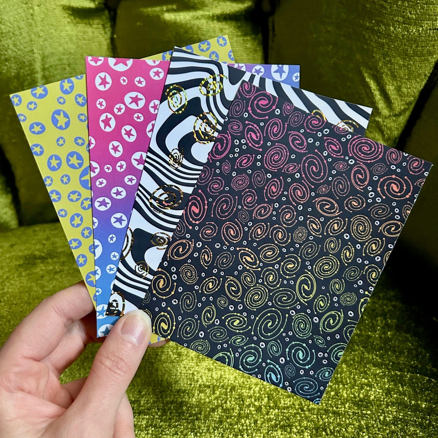 Four colorful patterned sticker sheets held by a hand against a green textured background