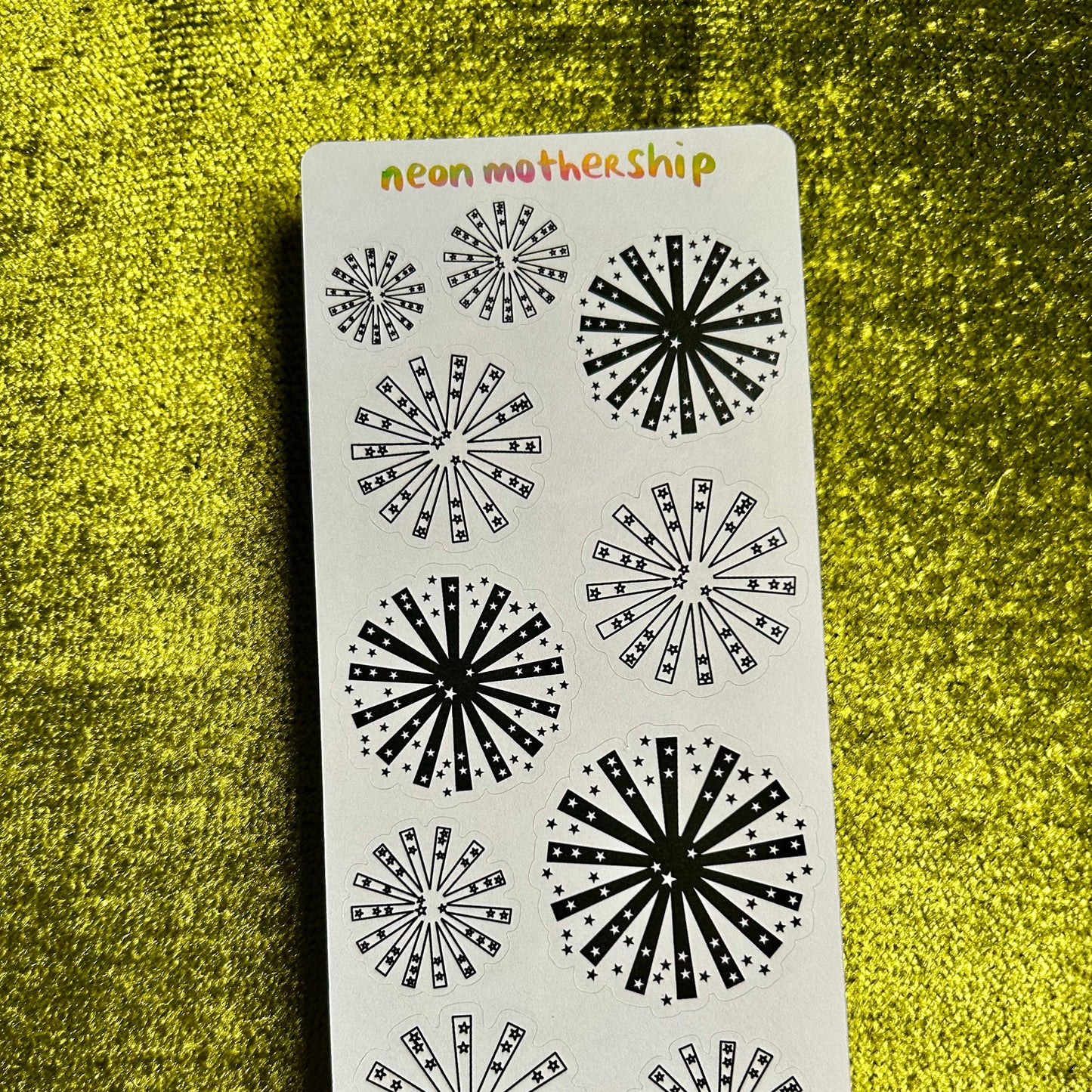 Sticker sheet with black starburst designs on a white background, labeled 'neon motherShip', against a green textured surface.