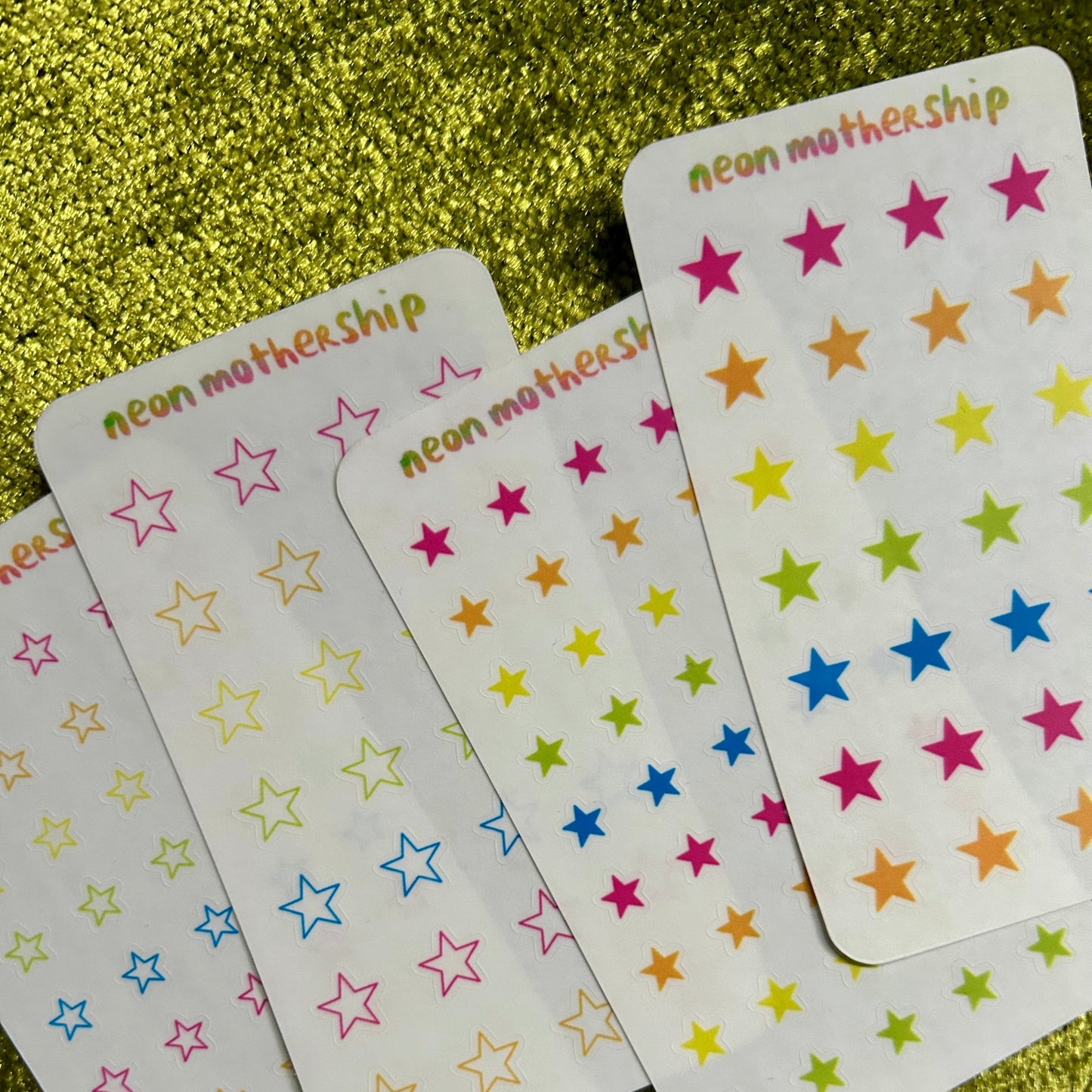 Colorful star stickers on white sticker sheets with 'neon mothership' branding on a glittery green background.