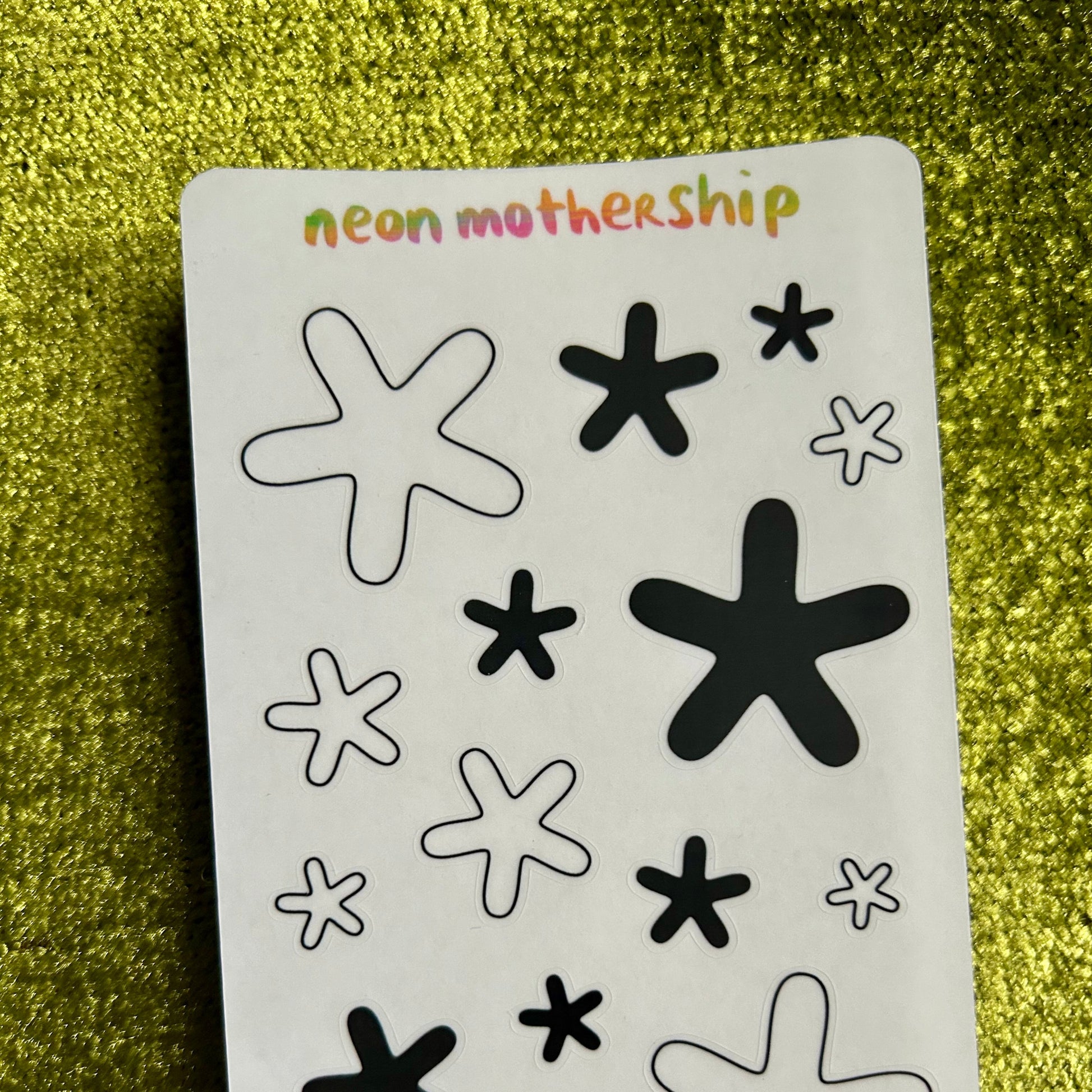 Sheet of black star-shaped stickers on a white card with 'neon mothership' branding, against a gold glittery background.