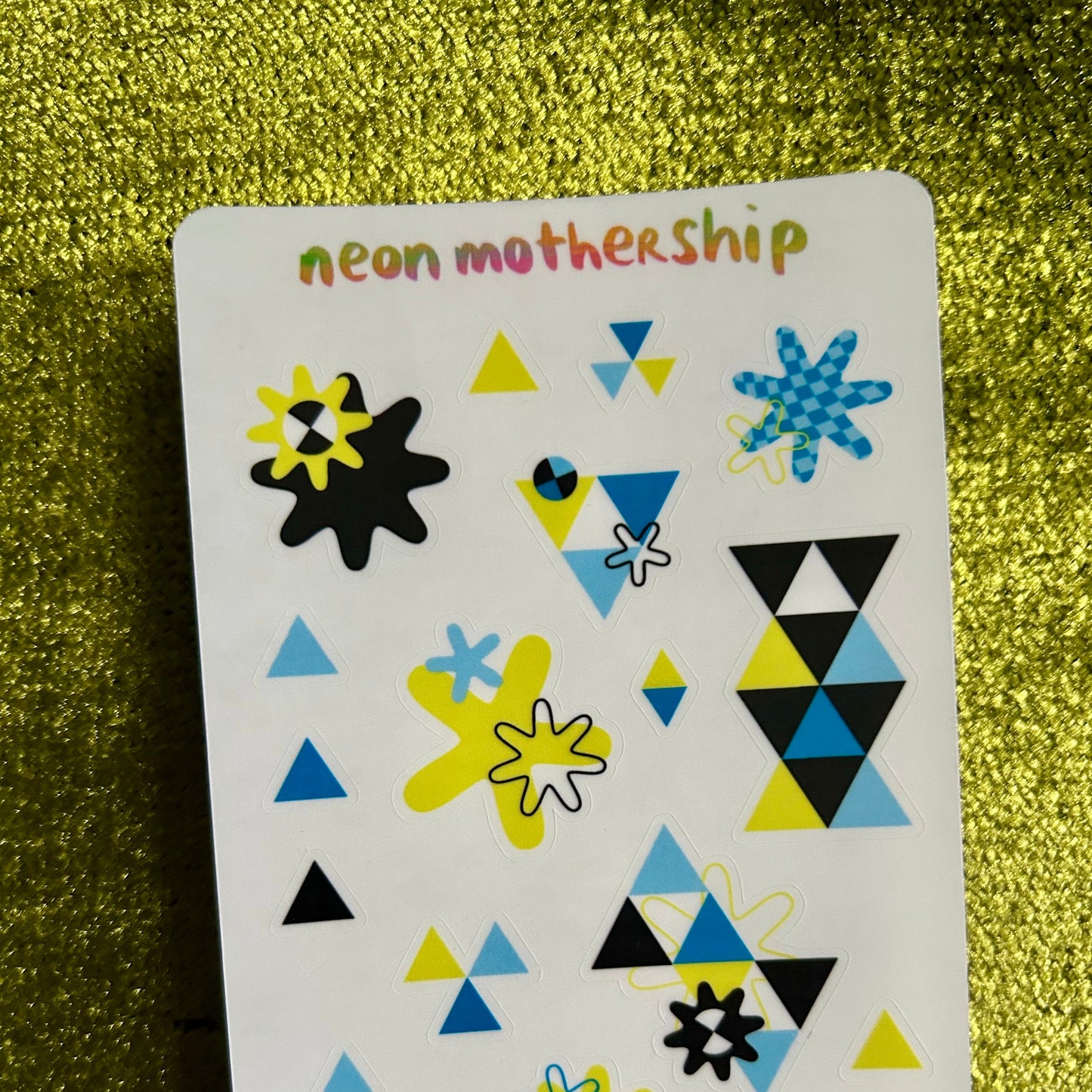 Sheet of geometric stickers with triangles, stars, and flowers on a green glittery background.