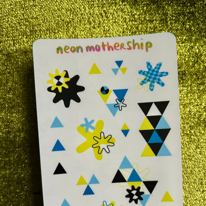 Sheet of geometric stickers with triangles, stars, and flowers on a green glittery background.