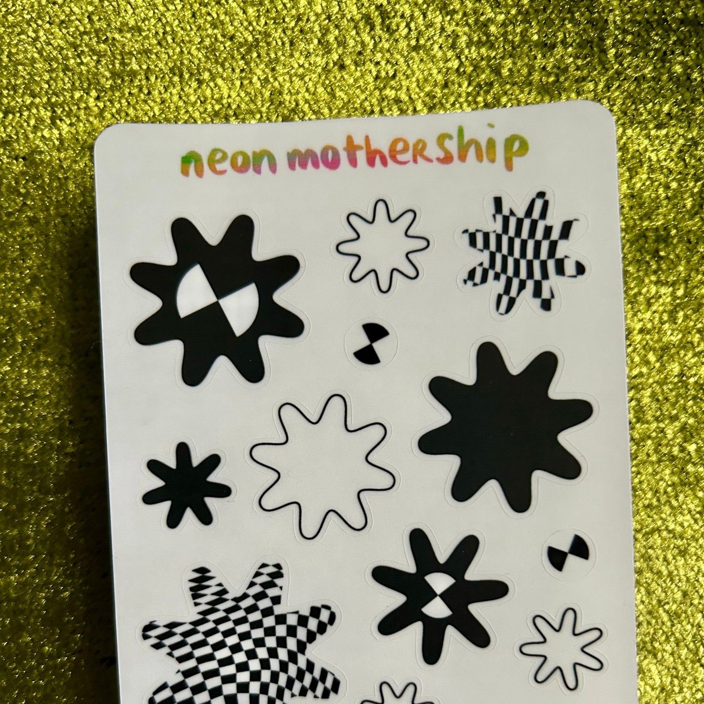 Sheet of black and white star-shaped stickers with 'neon mothership' branding on a gold glittery background.