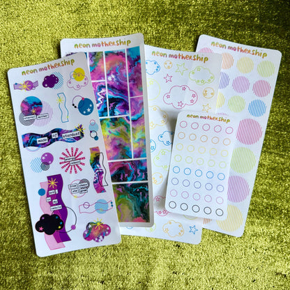 Colorful sticker sheets from 'neon motherShips' on a green textured surface