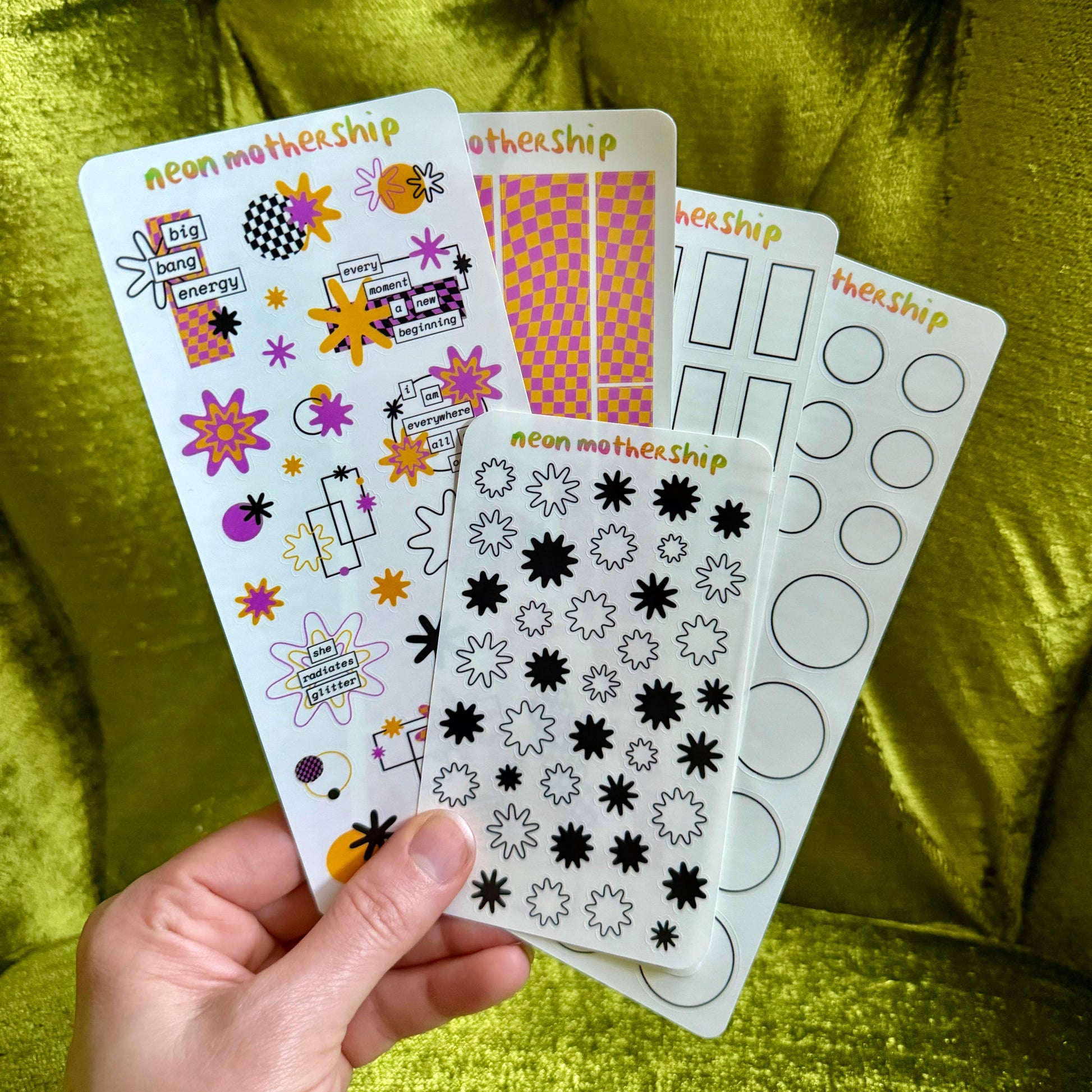 Hand holding a sheet of decorative stickers with various designs on a green fabric background