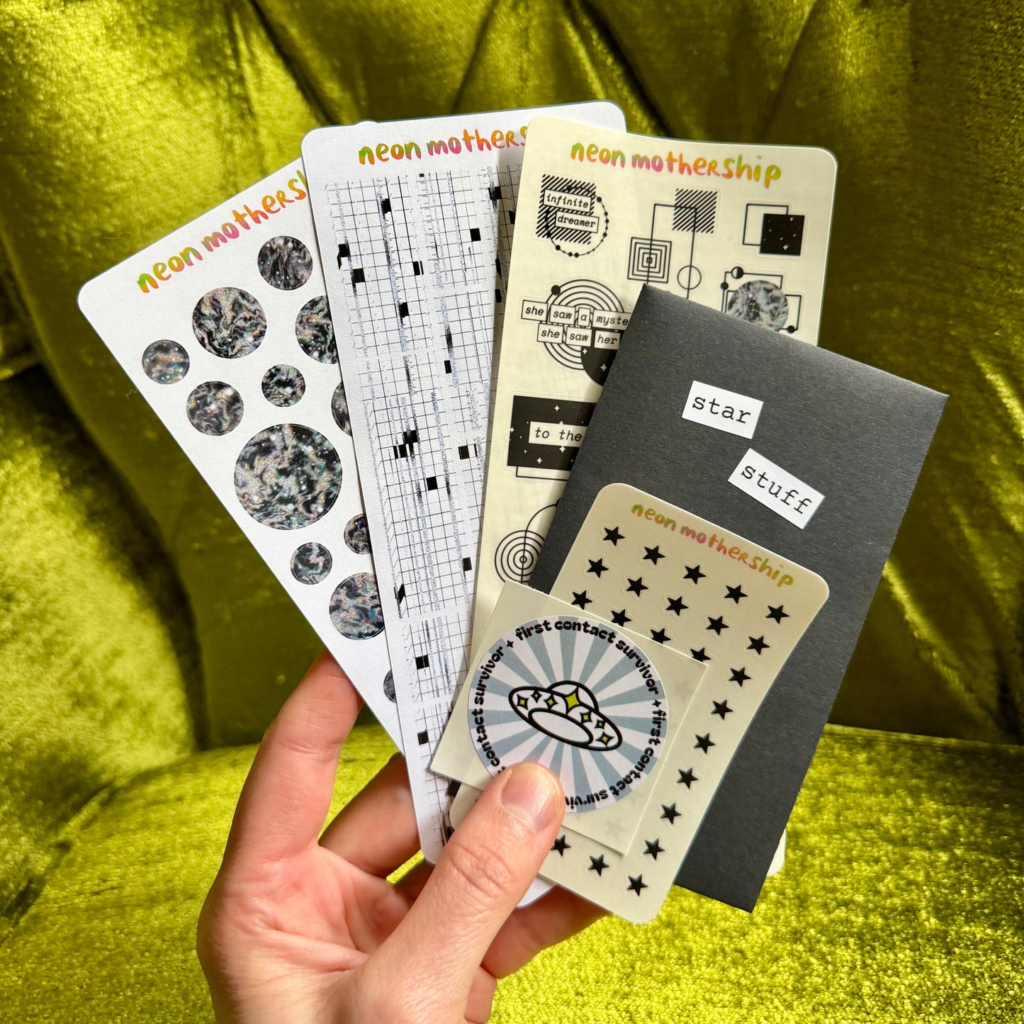 Hand holding a set of monochrome sticker sheets with 'neon mother ship' branding against a green textured background
