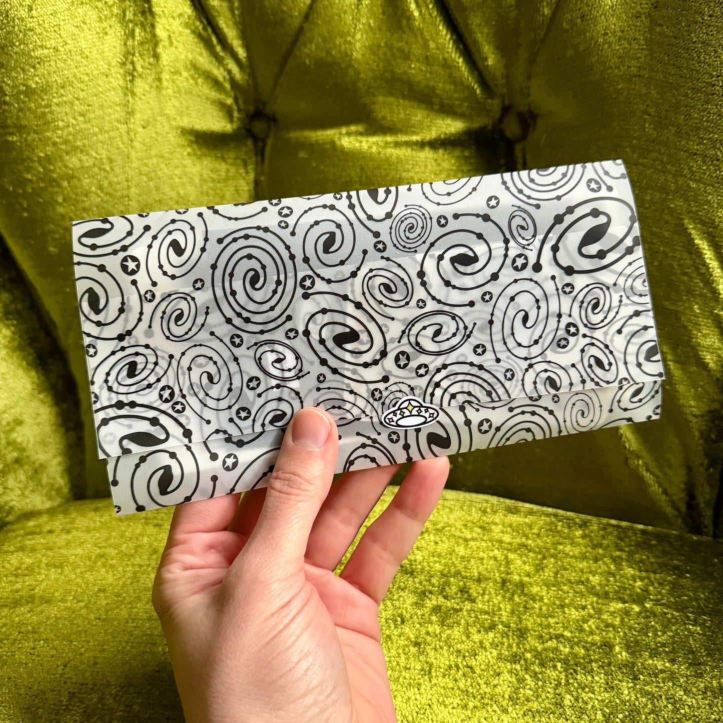 Hand holding a black and white patterned vellum envelope against a green fabric background