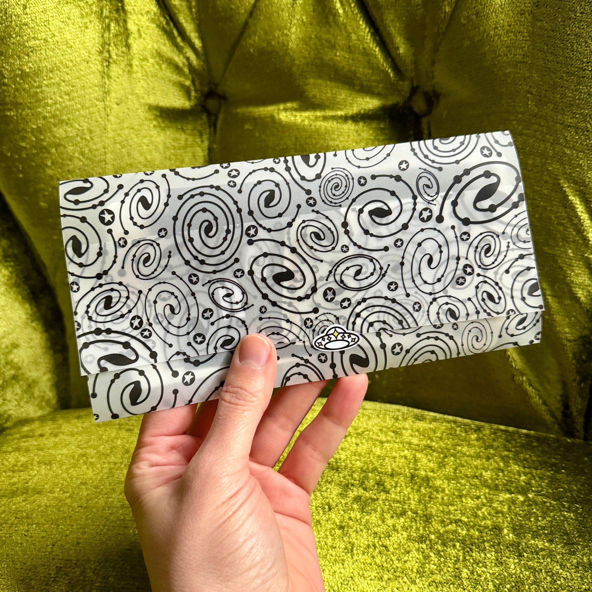 Hand holding a black and white patterned vellum envelope against a green fabric background