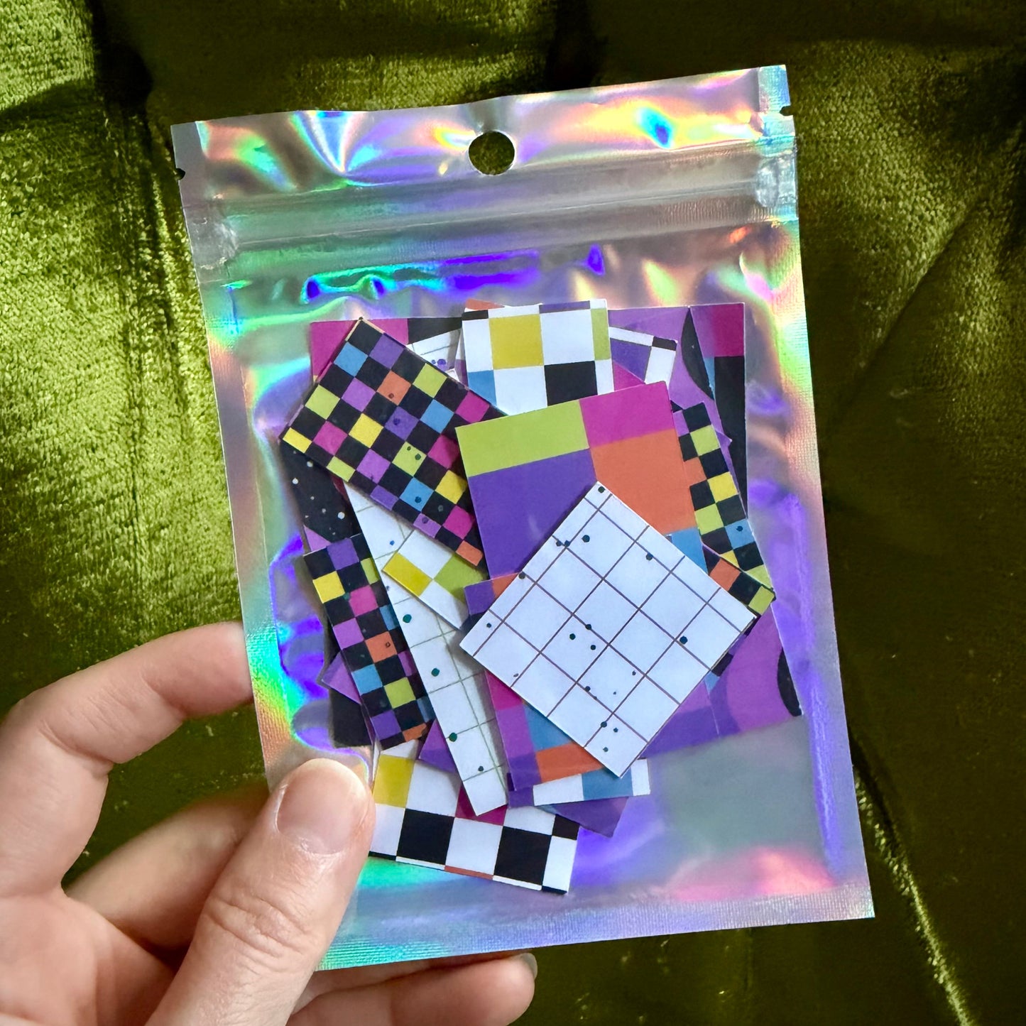 hand holding a small holographic package of colorful sticker scraps against a green background