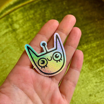 Holographic alien space cat sticker held in a hand with a green textured background