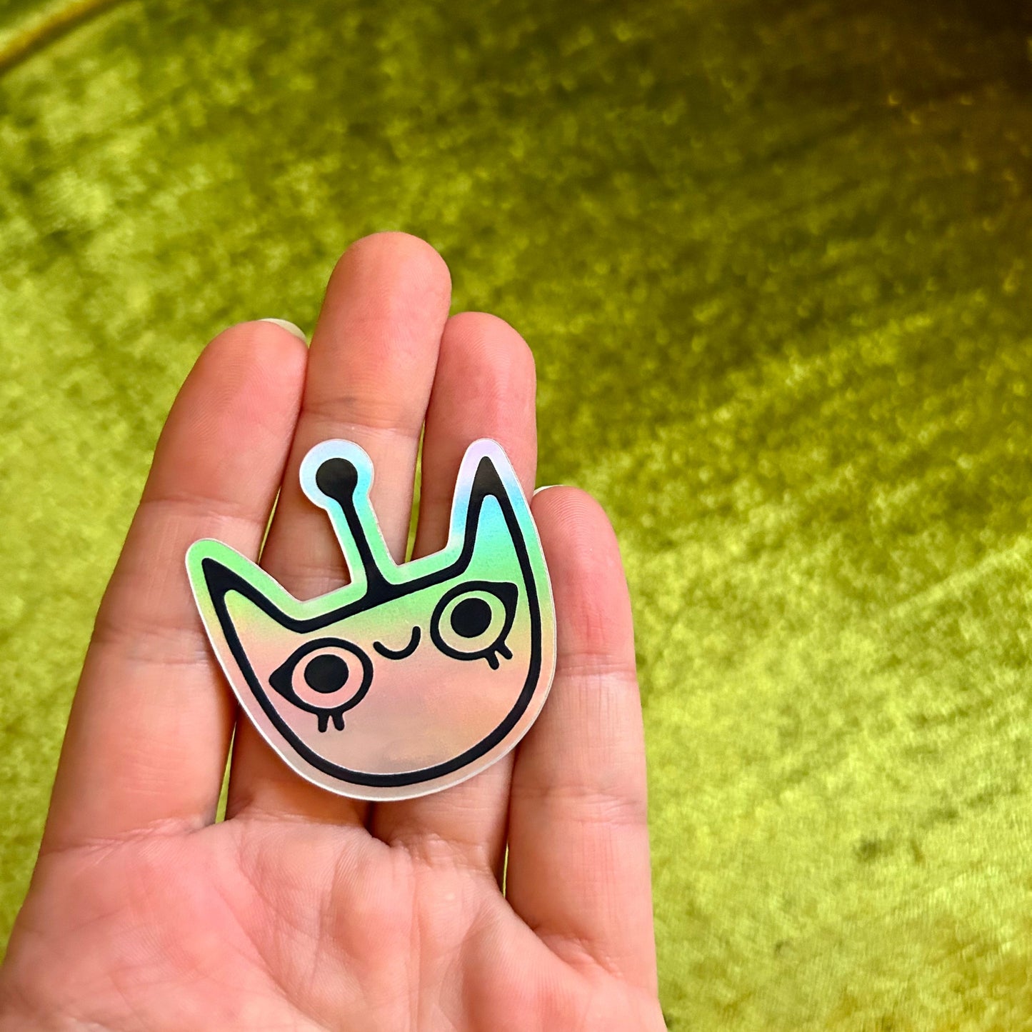 Hand holding a colorful alien face sticker against a green background
