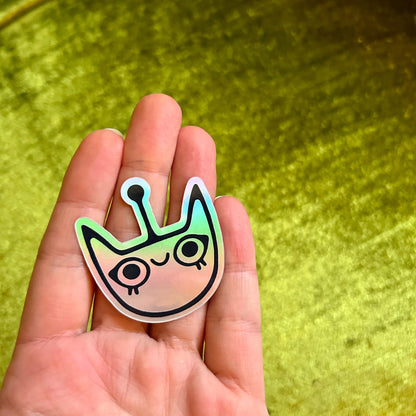 Hand holding a colorful alien face sticker against a green background
