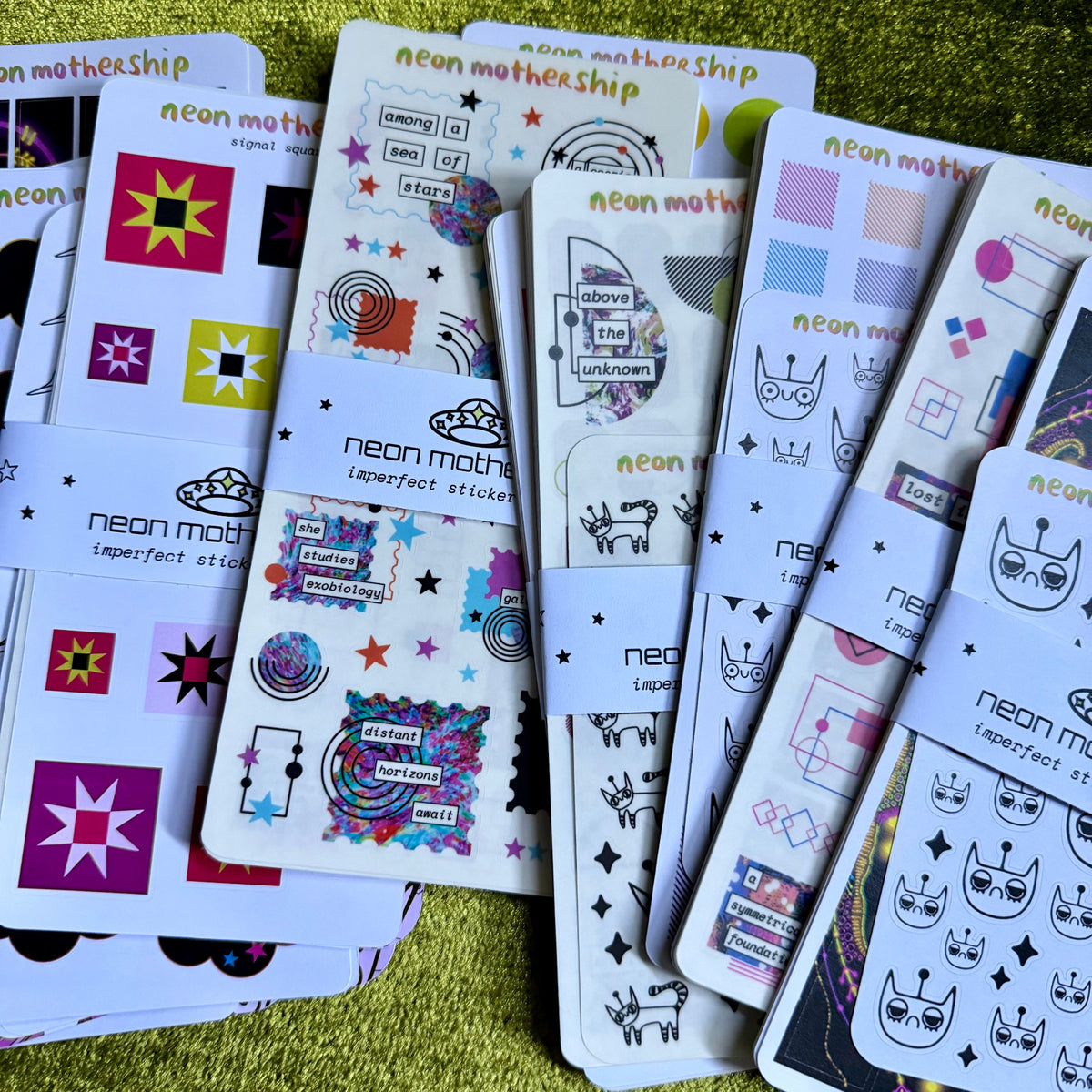 A fan of decorative sticker sheets with colorful neon-style graphics, stars, moons, and text such as 'neon mothership' and 'neon mothership imperfect stickers,' arranged on a textured yellow-green fabric background.