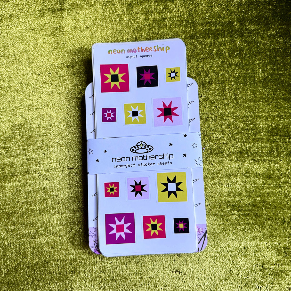 A stack of 'neon mothership' imperfect sticker sheets held together by a branded paper band, displayed on a textured olive green background.
