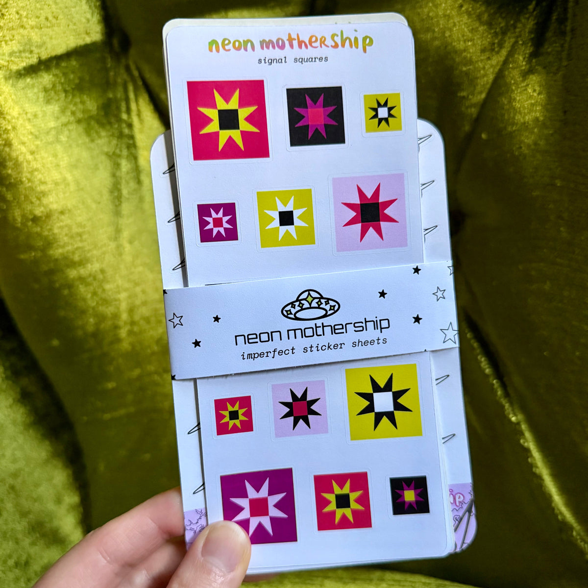 A hand holds a packaged sticker sheet set against a shiny olive green fabric background. The packaging is white with colorful geometric starburst designs and the brand name 'neon mothership' visible on the wrap.