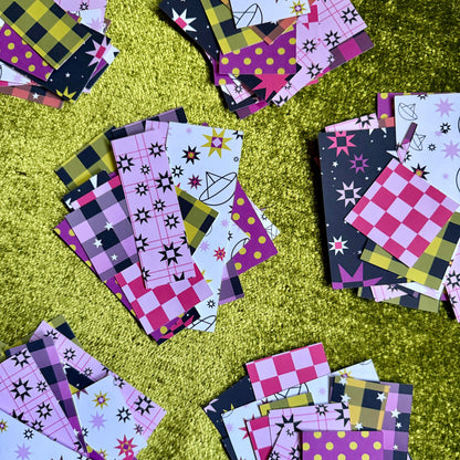 Multiple small patterned sticker paper pieces scattered on a shiny green surface. The pieces show pink, purple, black, yellow, and white patterns including plaid, checkers, polka dots, stars, and abstract shapes.