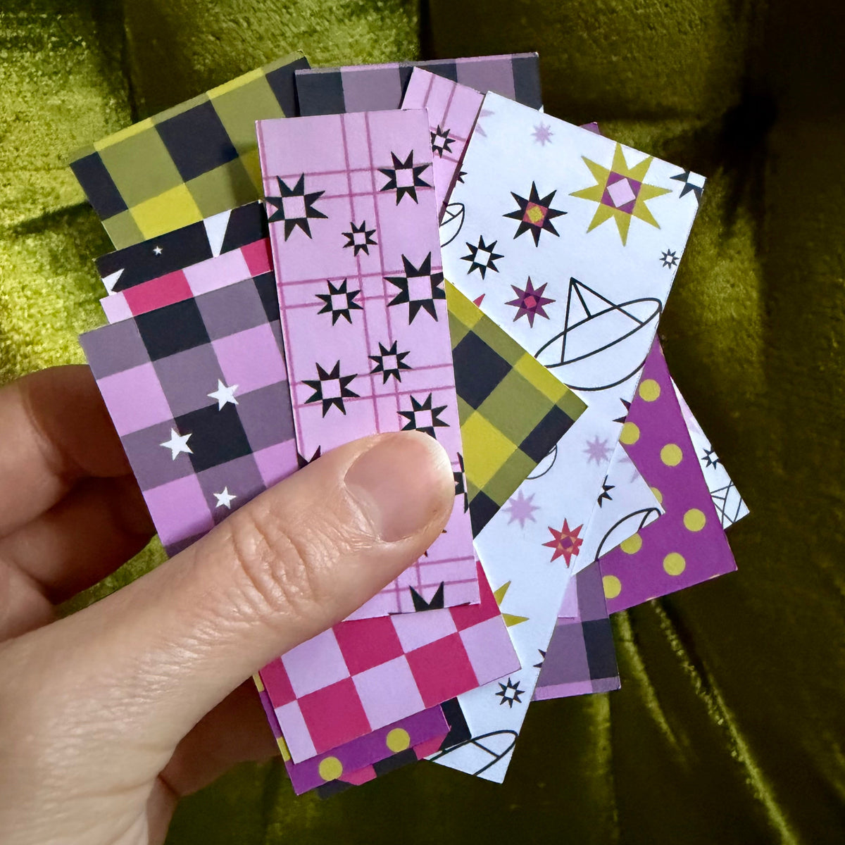 A hand holding a fan of small patterned pieces of sticker paper in pink, purple, yellow, black, and white, set against a shiny olive green velvet background.