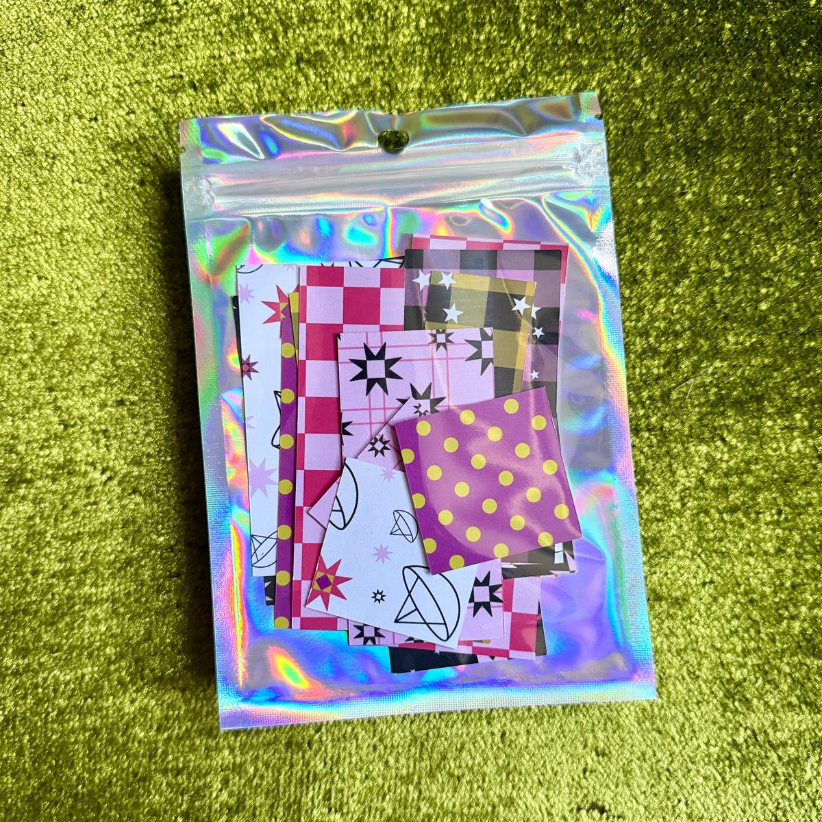 A small holographic zip pouch containing patterned sticker paper pieces, placed on a textured olive-green surface.