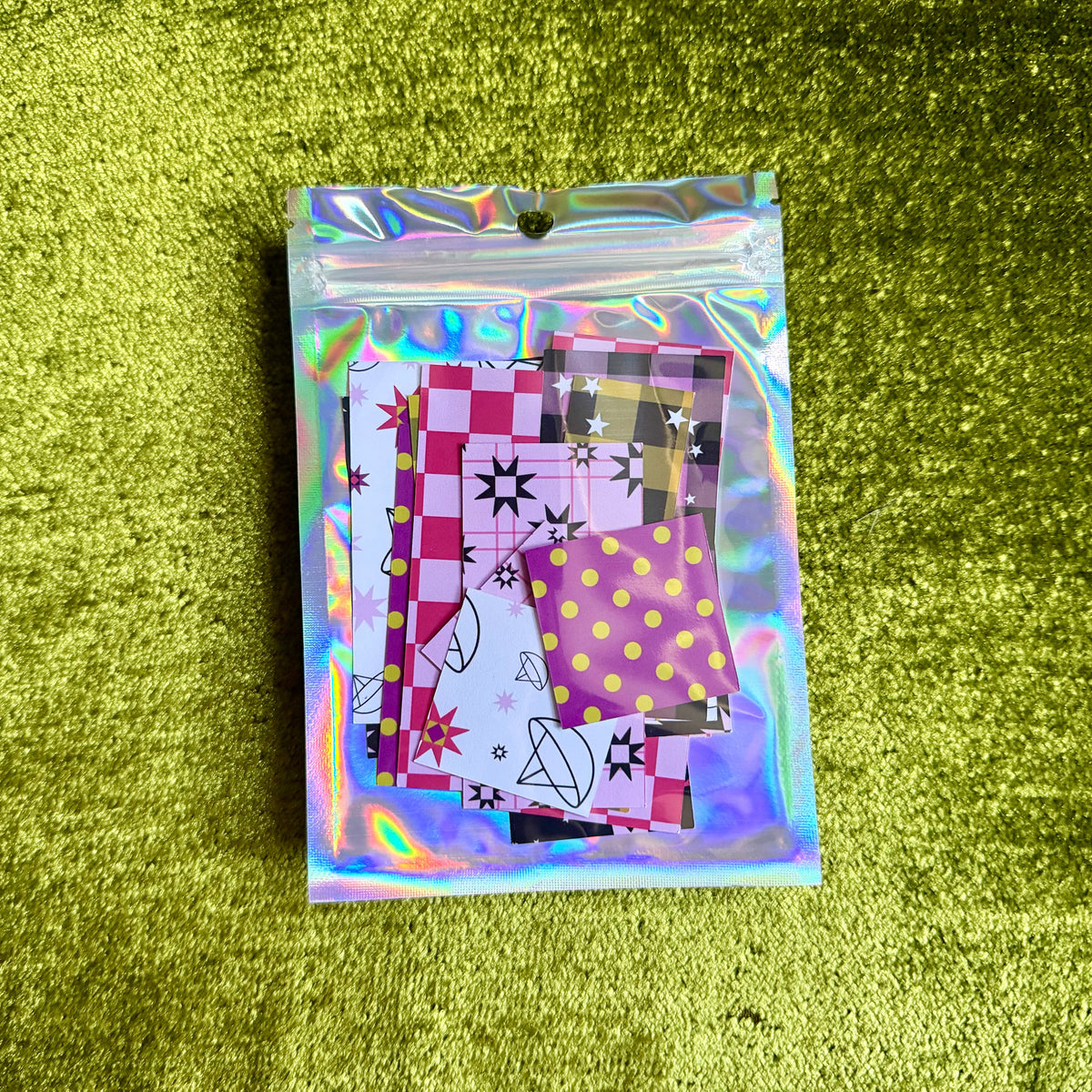 A small holographic zip pouch containing several patterned sticker papers placed on a textured olive-green surface.