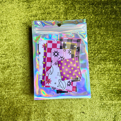 A small holographic zip pouch containing several patterned sticker papers placed on a textured olive-green surface.