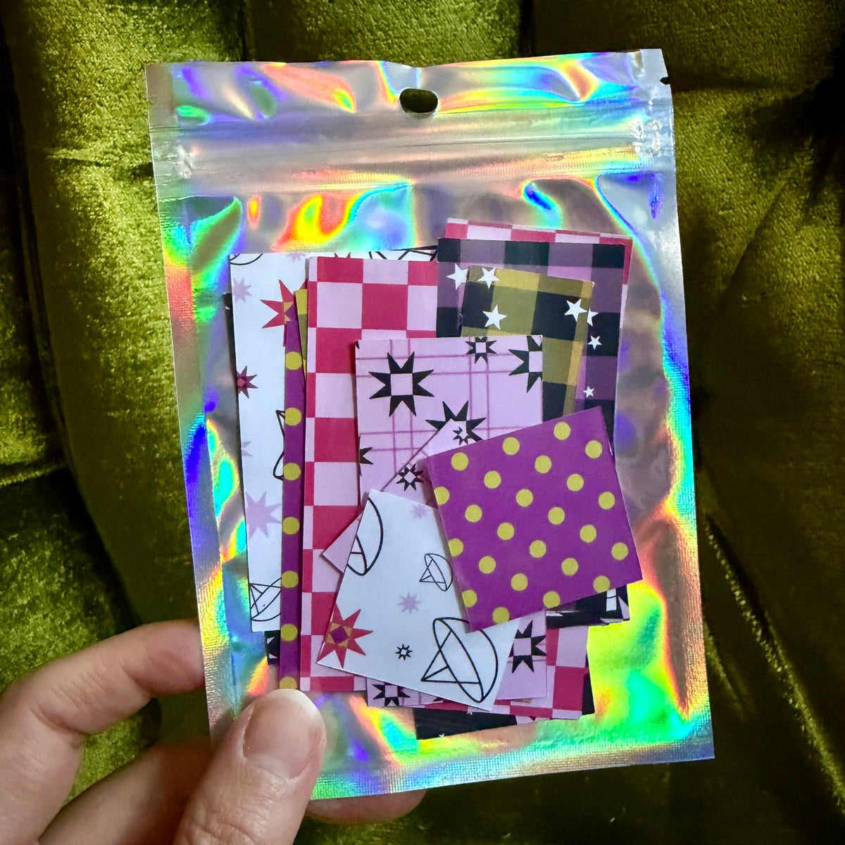 A hand holds a small holographic zip pouch containing several patterned sticker paper pieces with pink, purple, black, white, and yellow designs, set against a green velvet background.