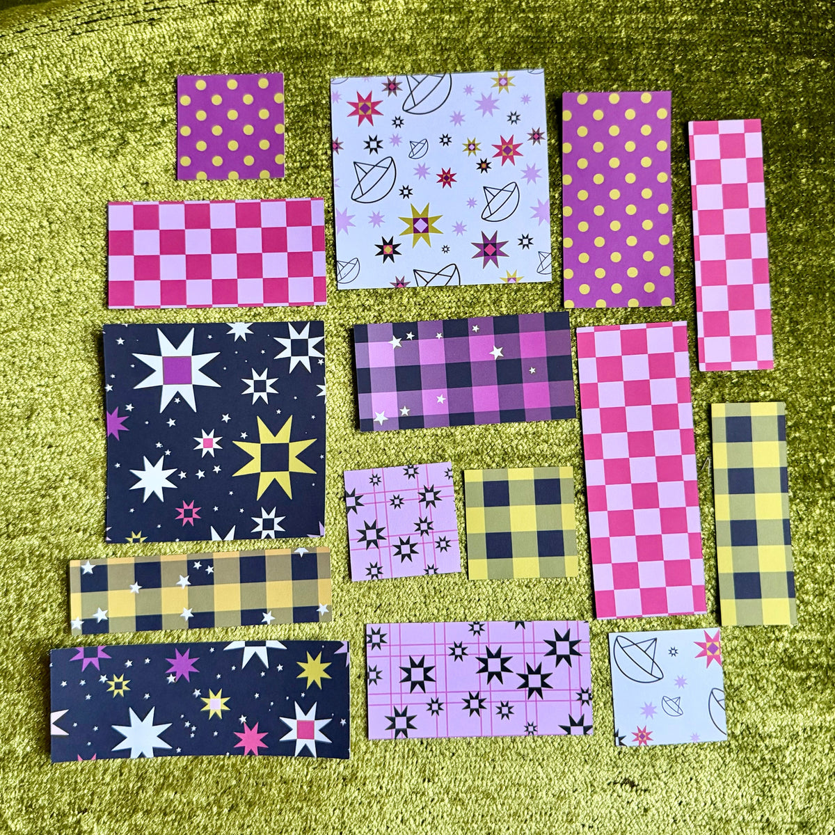 A collection of colorful patterned sticker scraps arranged on a textured olive green velvet surface. The pieces feature various patterns including pink checkers, yellow polka dots, black stars, purple plaid, and quilt star motifs.