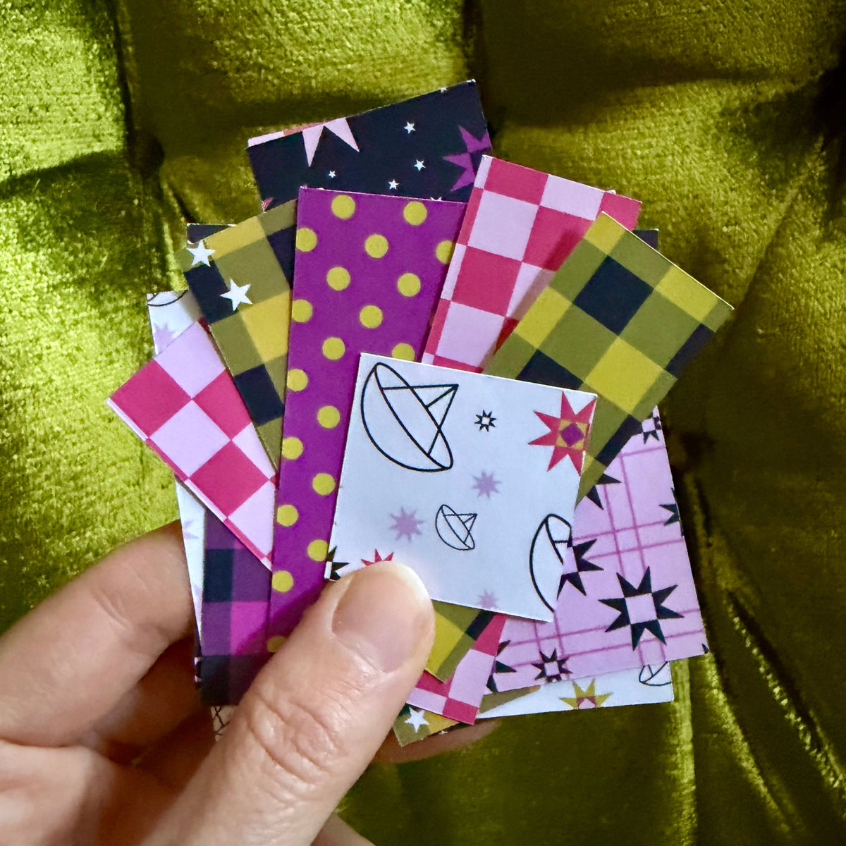 A hand holding a fanned-out selection of colorful patterned sticker paper squares, including plaid, polka dots, stars, and abstract designs, set against a shiny olive green fabric background.